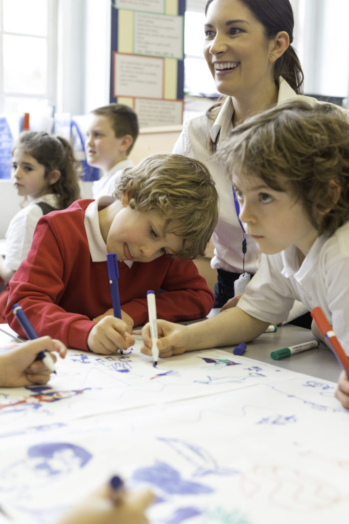 A small group of children are sitting around a desk with the teacher and drawing pictures and brainstorming on a large sheet of paper.