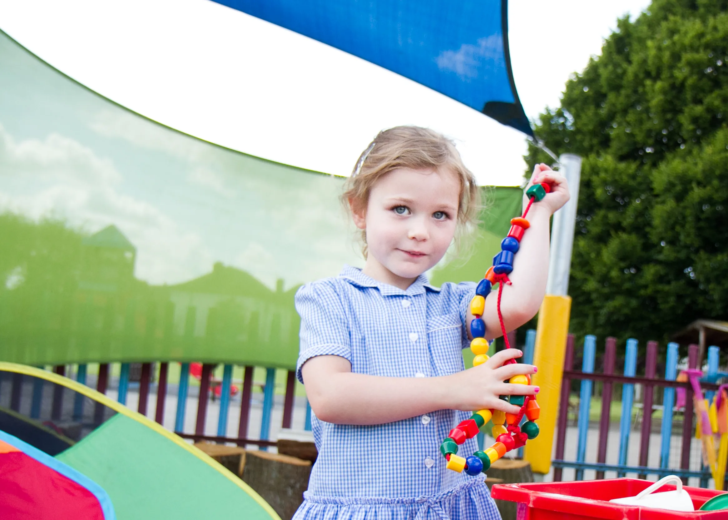 A young girl in a blue dress stands outside holding a colorful bead toy. She is surrounded by playground equipment and large fabric sunshades.