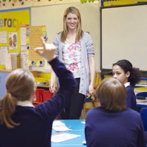 A classroom environment in a school. We see a teacher at the front of the class smiling at the students sat before her, who are engaged with her, and several pupils have their hands raised. It looks like a happy and nurturing learning environment.