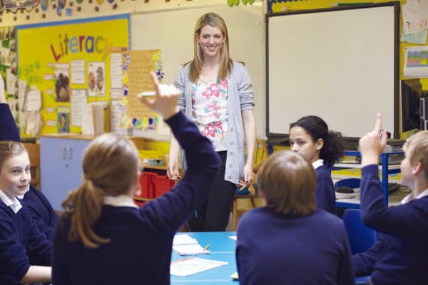 A classroom environment in a school. We see a teacher at the front of the class smiling at the students sat before her, who are engaged with her, and several pupils have their hands raised. It looks like a happy and nurturing learning environment.