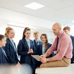 A group of secondary school pupils in a classroom are gathered round a teacher. The teacher is leaning on a desk, pointing at a paper he is holding and pleasantly explaining something to the pupils. They all look calm and happy, and nurtured.