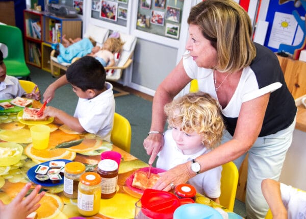 Nurture room pictured with children sitting at a table eating toast. A teacher is helping one child spread their toast.