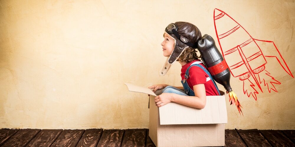 Small child sitting in a cardboard and wearing an helmet and backpack pretending to take part in a rocket launch